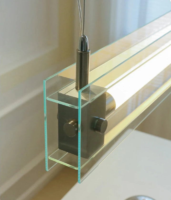 A modern, sleek linear glass pendant light with stainless steel accents, suspended over a counter, dispersing a warm glow.