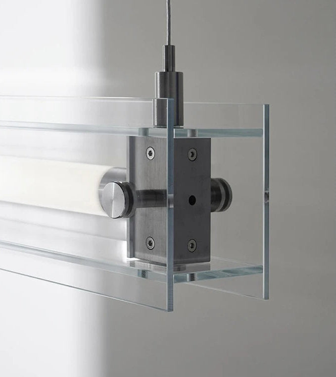 A modern, sleek linear glass pendant light with stainless steel accents, suspended over a counter, dispersing a warm glow.